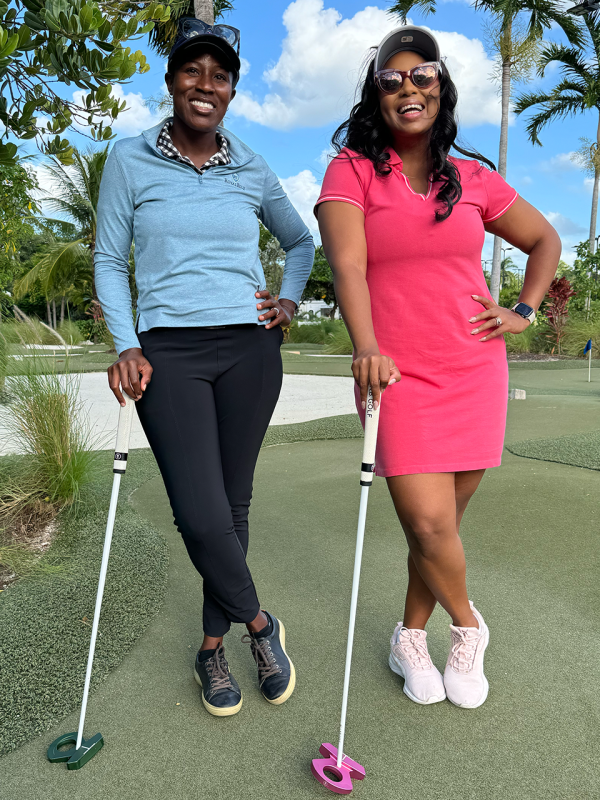 Two women stand beside each other leaning on golf putters.
